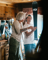 Två personer i en stuga som tittar på en Völkl Revolt 101 skida och diskuterar dess egenskaper.
