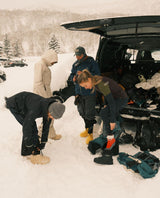 Skidåkare som tar på sig svarta och ljusa FUBUKI-vinterstövlar vid bakluckan på en bil i snöigt alpint landskap.