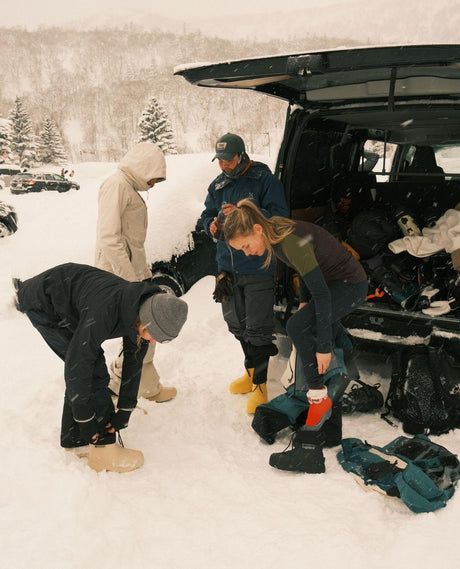 Skidåkare som tar på sig svarta och ljusa FUBUKI-vinterstövlar vid bakluckan på en bil i snöigt alpint landskap.