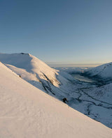 Skidåkare som gör en topptur i soligt alpint landskap med snöklädda berg och fjord i bakgrunden. Extrem Drifter 102.
