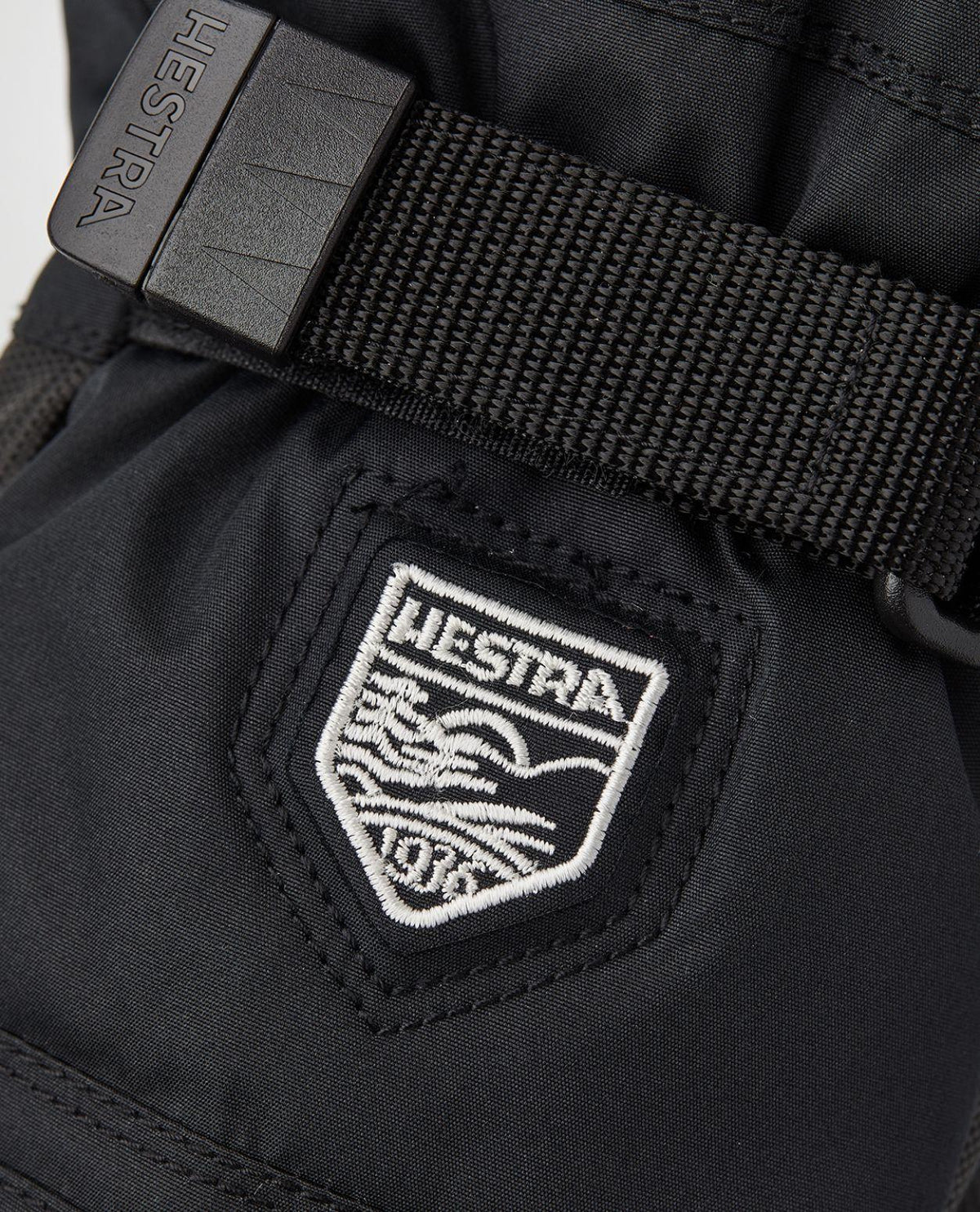 Hestra Gore-Tex Gauntlet Junior Mitt i svart visar en detaljbild på den broderade logotypen och handledsremmen.