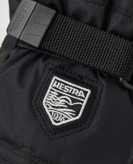 Hestra Gore-Tex Gauntlet Junior Mitt i svart visar en detaljbild på den broderade logotypen och handledsremmen.