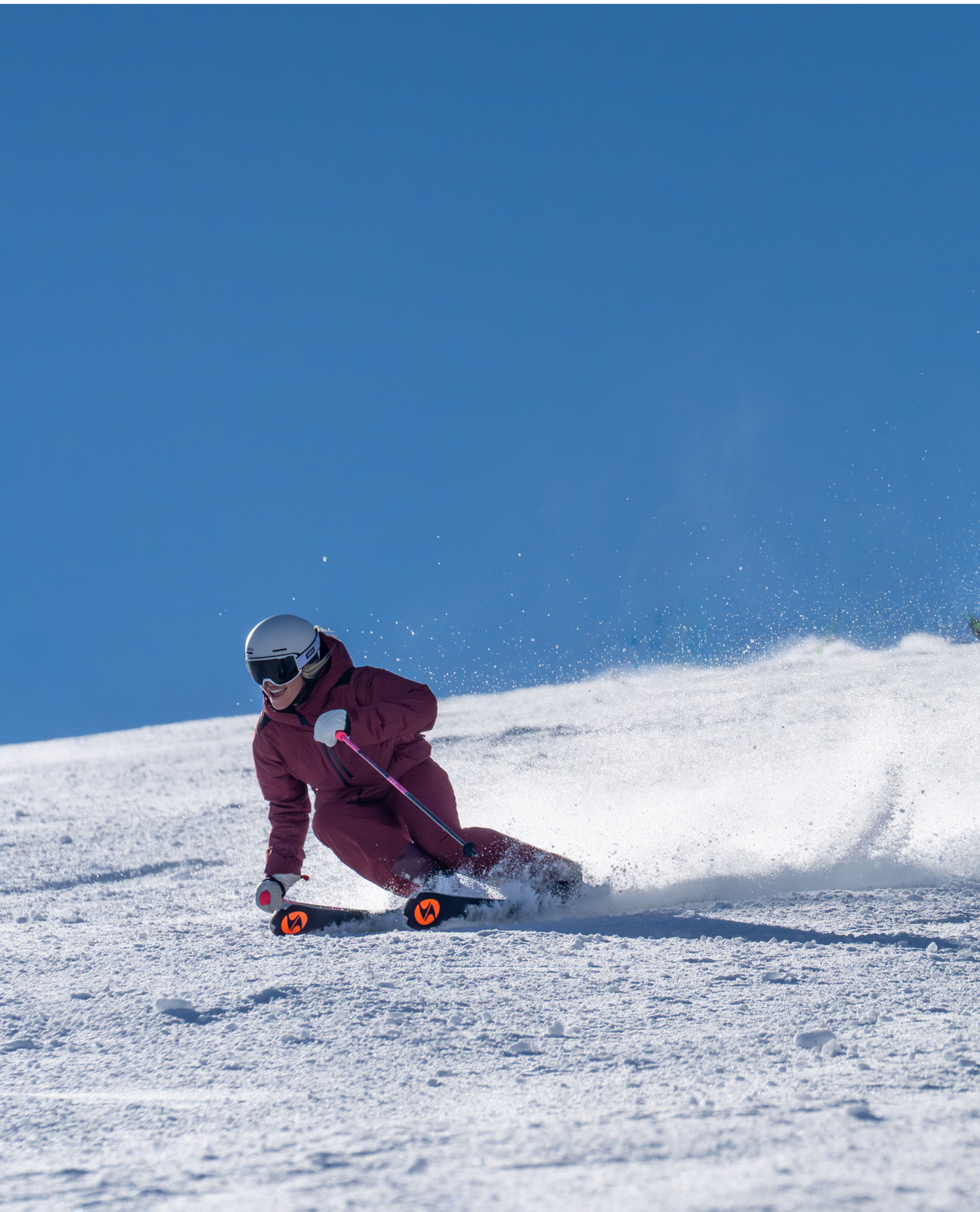Kvinna i röd skidoutfit som carving-svänger i pisten med Blizzard Phoenix-skidor i strålande sol.