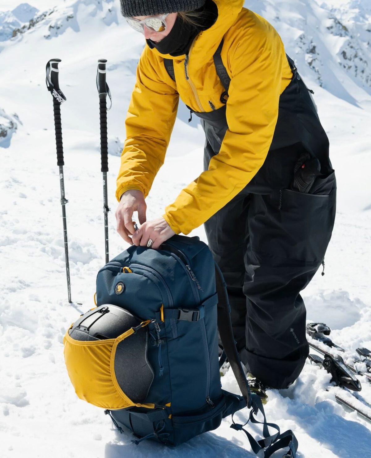 Skidåkare i svarta Bergtagen Gore-Tex Touring Bibs och gul jacka som packar ryggsäck i snölandskap.