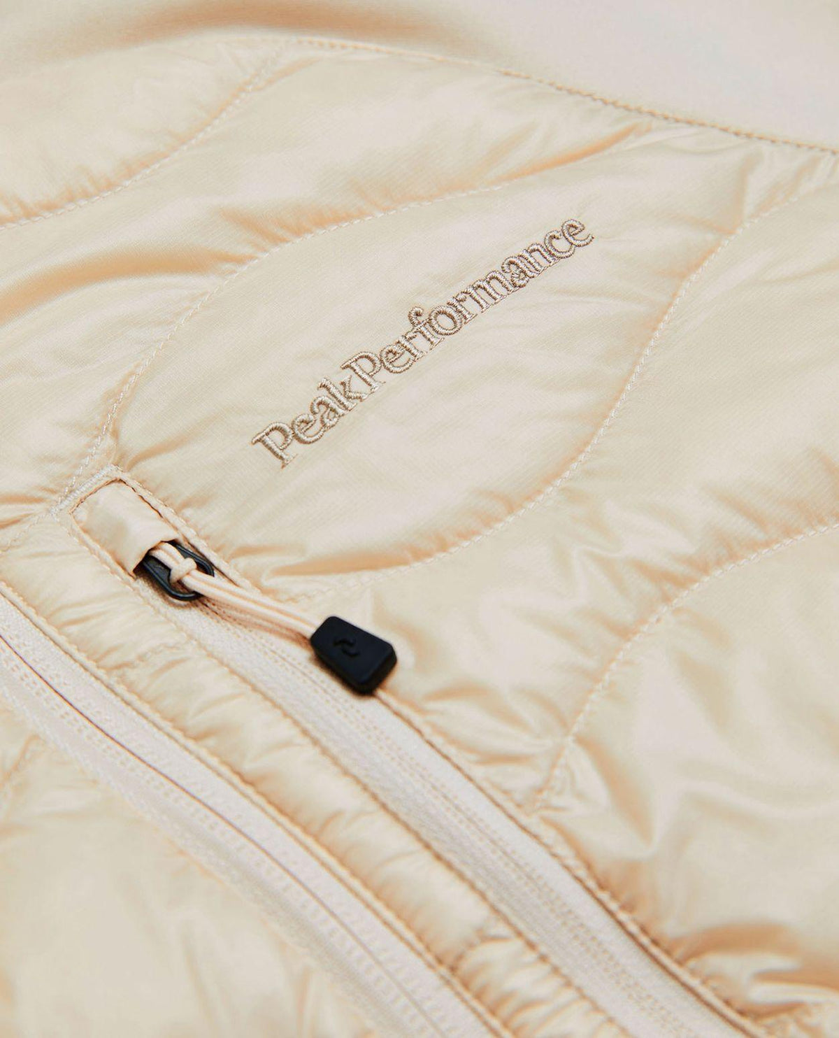 Närbild på Peak Performance Helium Down Hybrid Hood för damer i färgen sand fog med broderad logotyp och dragkedja.