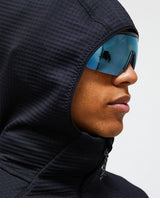 Närbild i profil på en skidåkare i svart huvtröja med luvan uppdragen och blå spegelglasögon.