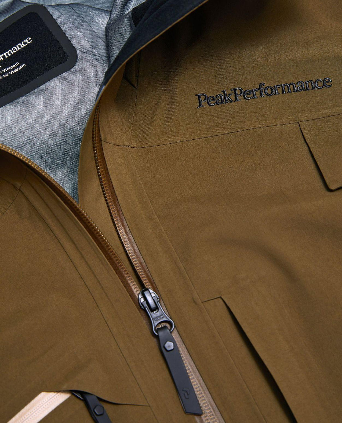 Närbild på broderad logotyp och dragkedja på Peak Performance Alpine Gore-Tex 3L Jacket i färgen Meadow Moss.