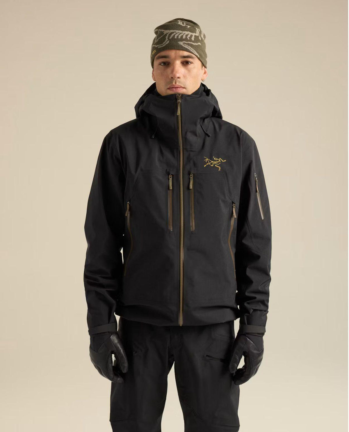 Man iklädd svart Arc'teryx skidjacka med luva, bröstfickor och synliga dragkedjor framifrån.