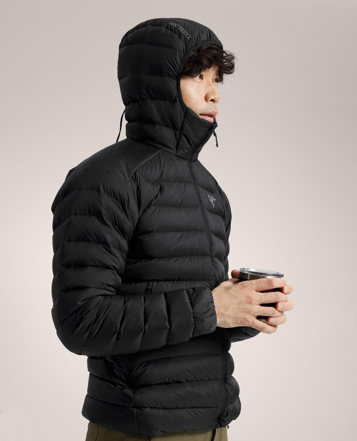 Manlig skidåkare i profil iförd den svarta dunjackan Arc'teryx Cerium Hoody med uppdragen huva och kopp i handen.