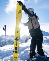 En person som gör en topptur håller upp ett par gula DPS Pagoda Tour CFL 112 25/26 i solen på en bergstopp - Alpingaraget