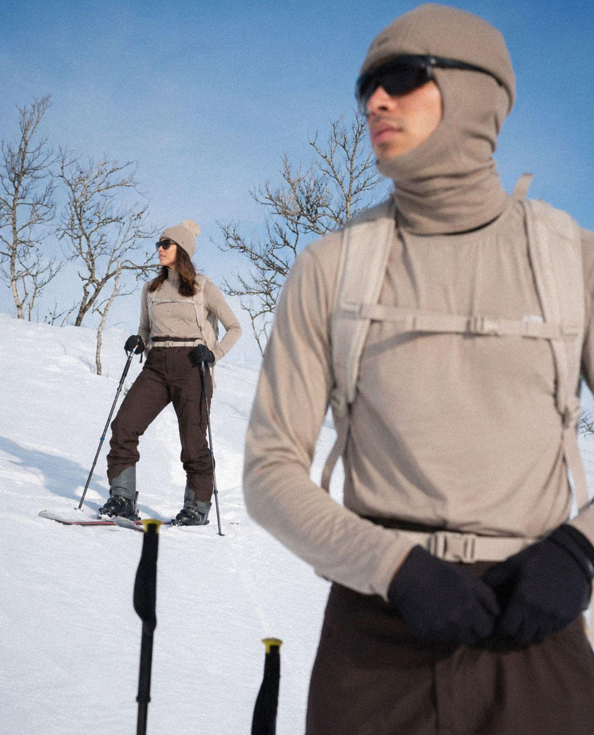 Skidåkare i beige tröja med huva och bruna skidbyxor som står i ett soligt snölandskap.