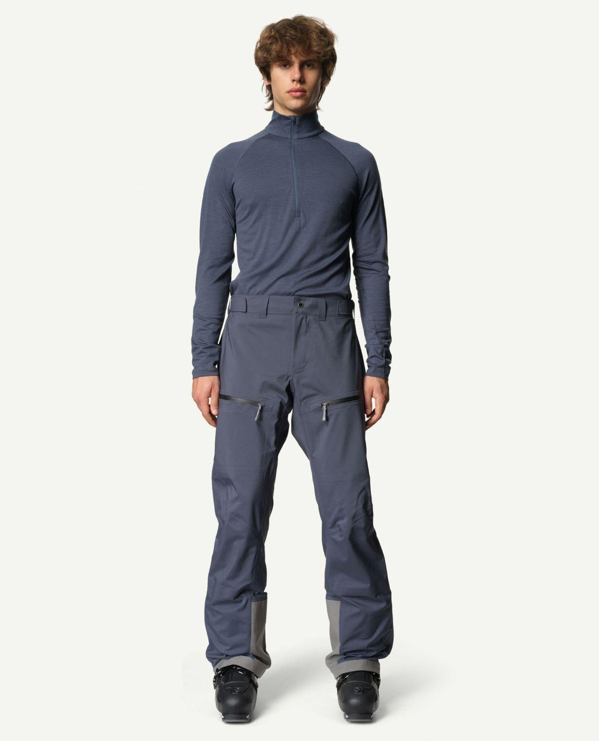 Manlig skidåkare iförd blå Houdini Ride Pants II skidbyxor och pjäxor framifrån.
